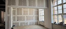 Renovierungsbaustelle mit verspachtelten Gipskartonwänden, Laserabstandsmessgerät auf Stativ und großem Bogenfenster.
